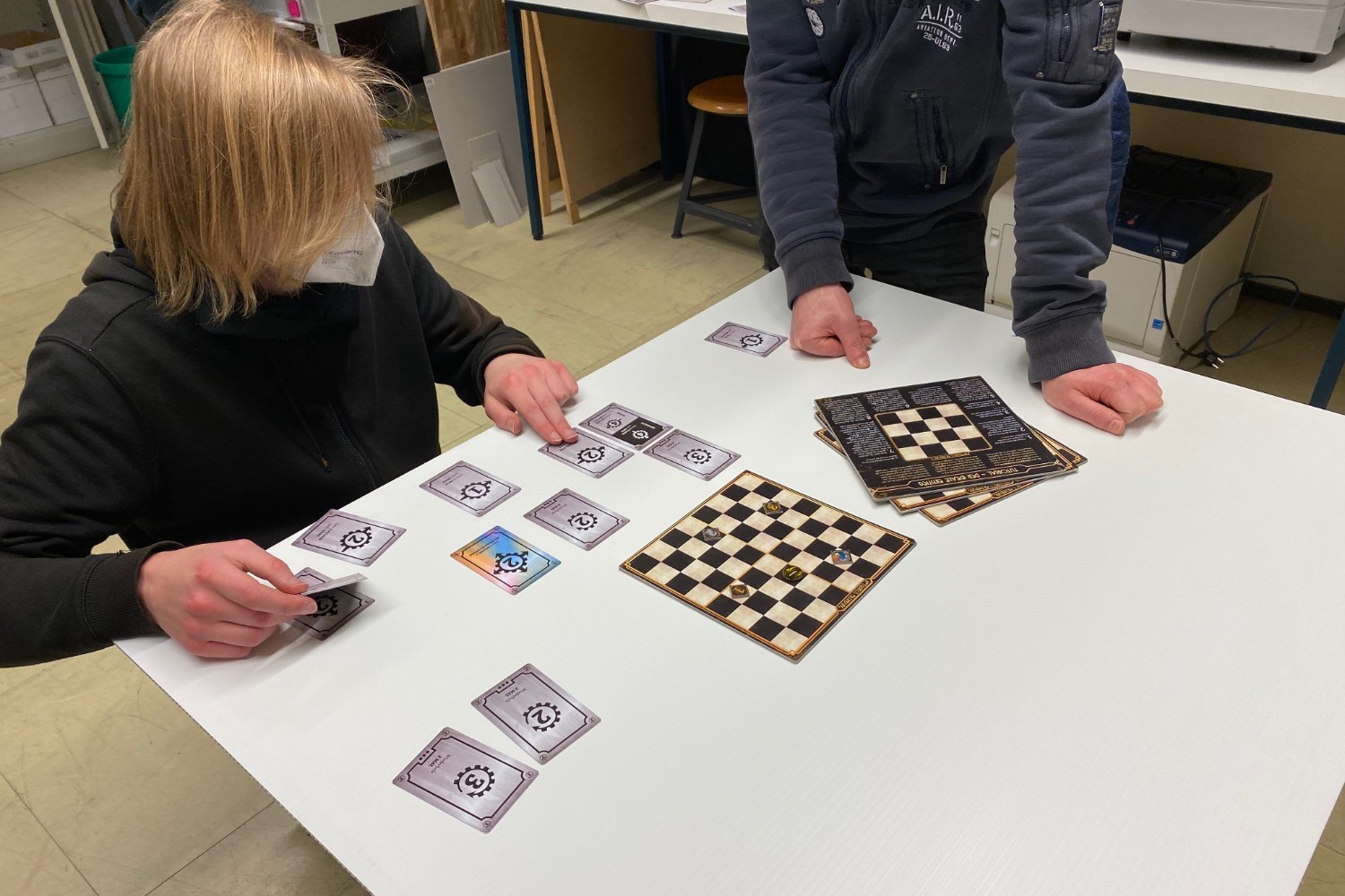 The game materials for “Rotwangs Erfindung” are laid out on a table and examined by two people.
