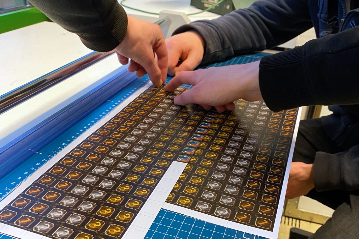 A printed sheet with tokens printed on it lies on a paper cutting board. Several people's hands touch the printed sheet